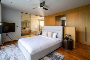 A modern bedroom with a large bed, a ceiling fan, a TV, and a bathroom visible through a glass door. - Villa Kaya (Thep Krasattri)