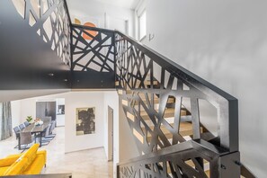 Interior moderno com escada com guarda-corrimão preto geométrico, área de refeições com uma mesa comprida e cadeiras amarelas, e cozinha ao fundo.
