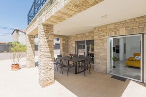 A covered patio with a stone wall, a dining table with chairs, and a sliding glass door leading to an interior space. - Villa Mirela - Perfect Family escape With  Private Pool, BBQ & Fenced Yard (Šišan)
