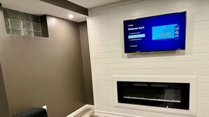 Ett modernt vardagsrum med en platt-TV som hänger ovanför en öppen spis, en soffa i mörk färg och ett trägolv.