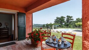 En overdækket terrasse med et bord dækket til to personer, en potteplante og udsigt til en swimmingpool og grøn bevoksning.