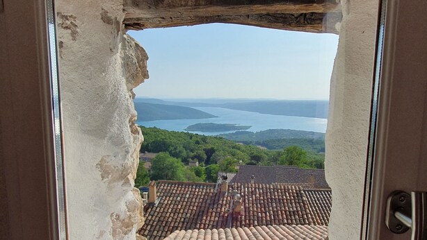 Janela com moldura danificada, vista para um telhado de cerâmica e um corpo d'água ao longe.