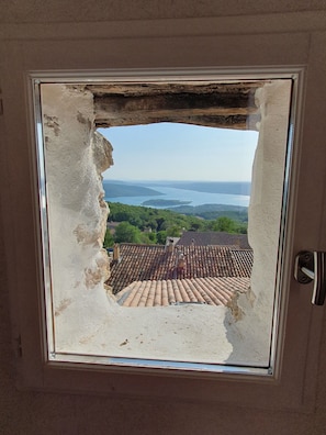 A window with a damaged frame, overlooking a tiled roof and a distant body of water.