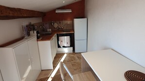Cozinha compacta com bancada branca, forno embutido, frigorífico e um banco de madeira.