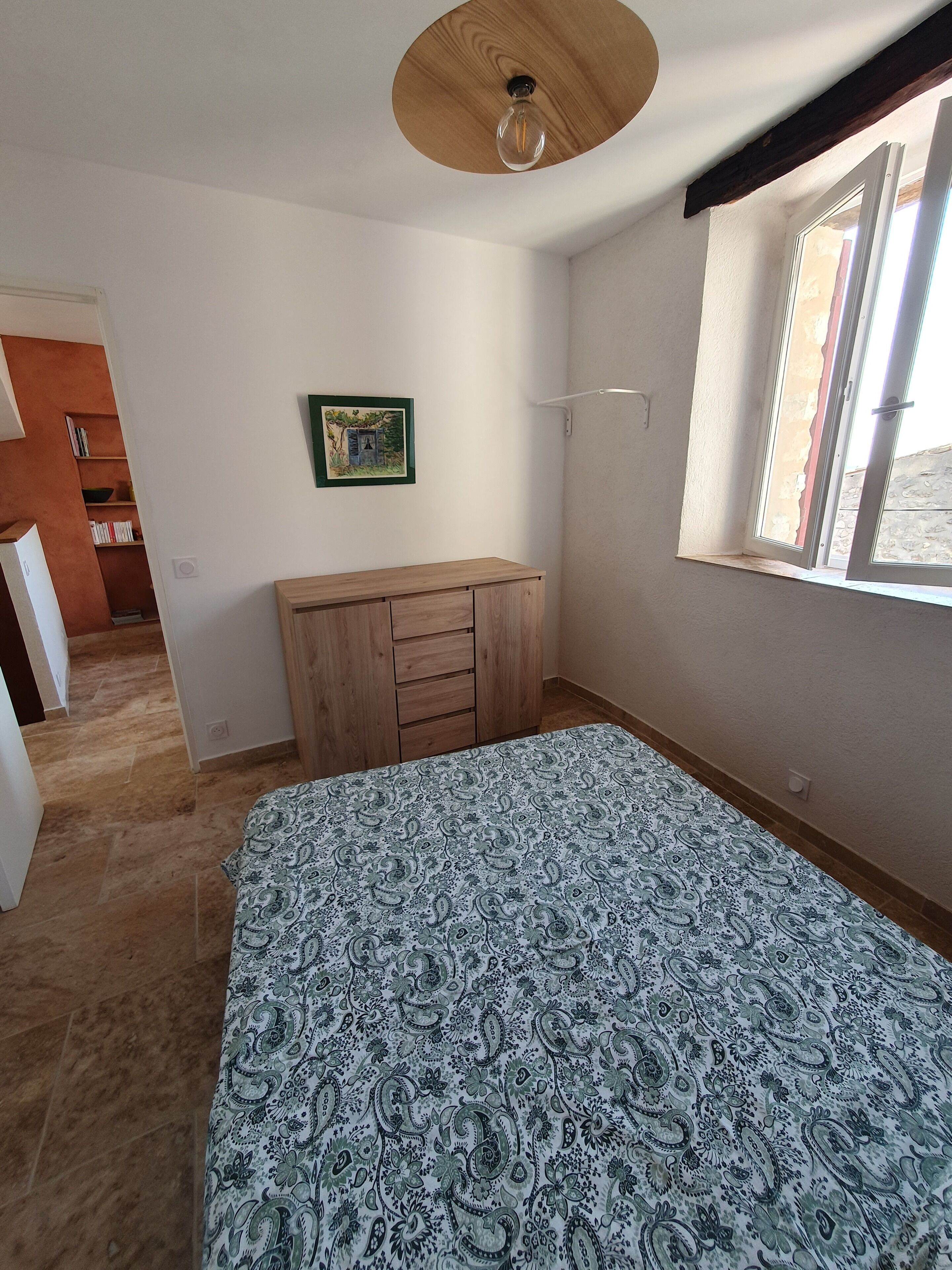 Une chambre à coucher avec un lit, une commode en bois, un tableau au mur et une fenêtre avec des stores.