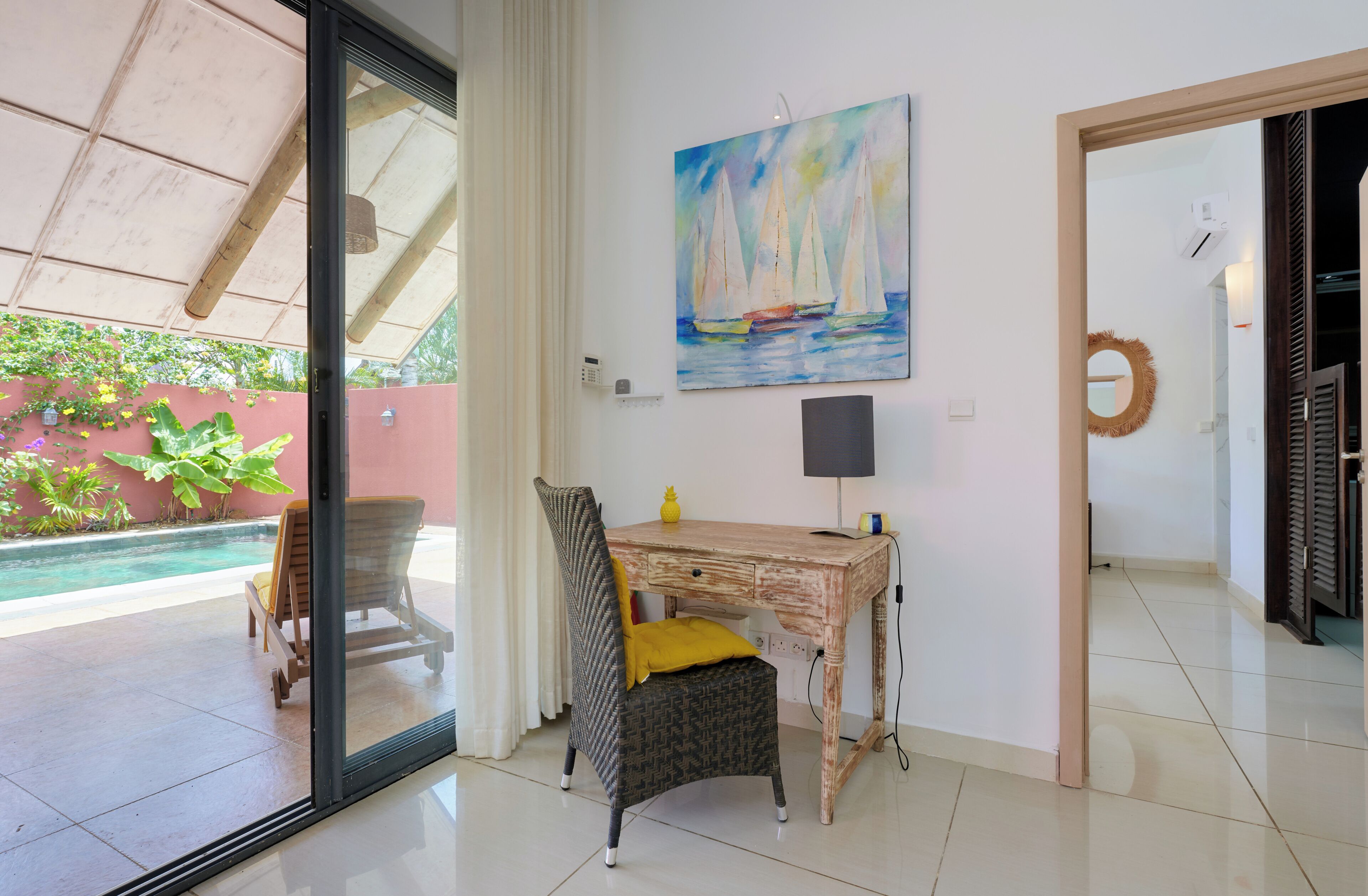 A room with a glass door leading to a pool, a wooden desk, a chair, and a painting of sailboats.