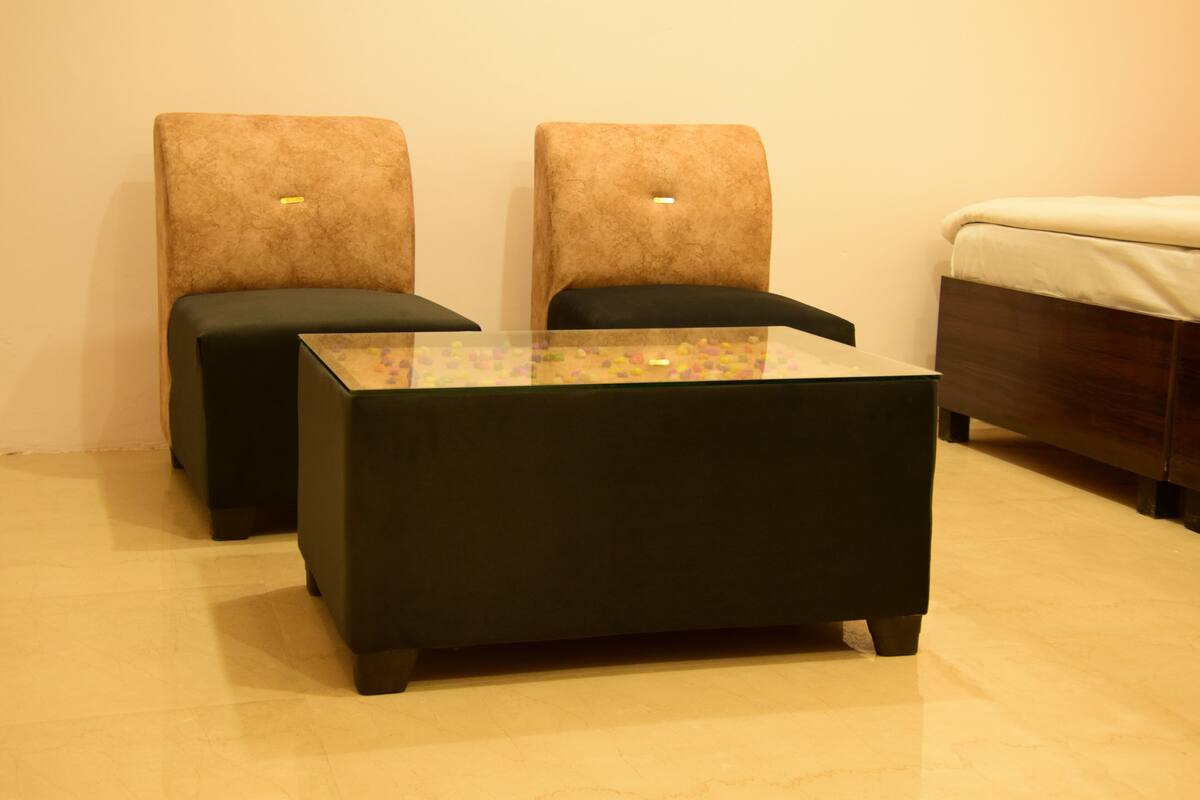 A room with a glass-top coffee table, two black ottomans, a bed with a wooden headboard, and a plain wall.