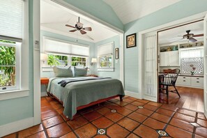 Ein Schlafzimmer mit einem Bett, einem Deckenventilator, einem Fenster mit Jalousien, einem Sessel und einer Küchenzeile mit Schränken und einem Fenster.
