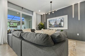 En moderne stue med en stor grå hjørnesofa, et indrammet billede på væggen og et stort vindue med gardiner.