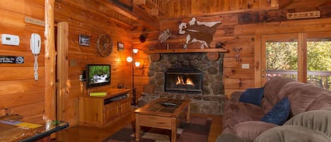 Ein Blockhaushaus-Interieur mit Kamin aus Stein, Holzmöbeln, einem Fernseher und einem Deckenventilator.