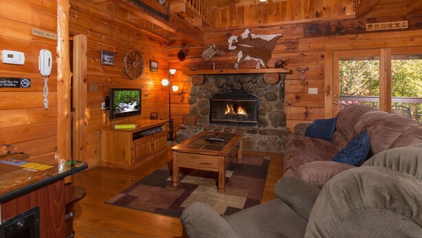 L’intérieur d’une cabane en rondins, avec une cheminée en pierre, des meubles en bois, un téléviseur et un ventilateur de plafond.