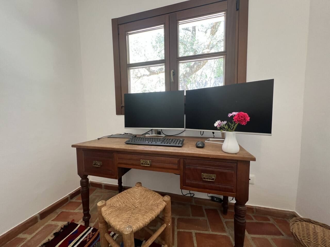Un escritorio de madera con dos monitores, un teclado, un ratón y una jarra con flores.