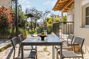 An outdoor patio with a table and chairs, surrounded by plants and a view of a building. - Spacious villa with beautiful large garden (NEA MOUDANIA)