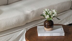 En sofa med pude, et rundt træbord med en bog og en vase med blomster.
