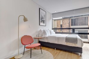 A modern bedroom with a bed, a red chair, a floor lamp, a small table, and a large window with a view of a brick building. - w Premium Loft in the Heart of Bogot (Bogotá)