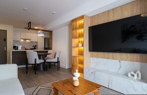 Een moderne woonkamer met een houten eettafel, een bank, een aan de muur bevestigde televisie en een keuken op de achtergrond.