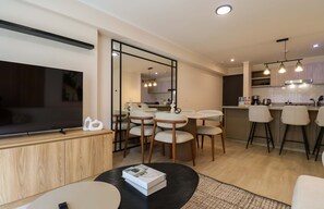 Ein modernes Wohnzimmer mit Flachbildfernseher, ein Essbereich mit rundem Tisch und Stühlen sowie eine Küche mit Barhocker und Pendelleuchten.