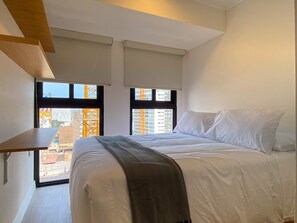 Quarto com cama, duas janelas, vista para prédios e teto de madeira.