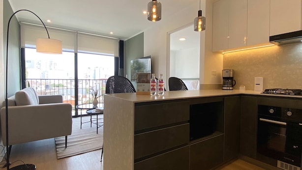 A modern kitchen with a built-in stove, a coffee maker, and a dining area with a sofa and a table. - w Deluxe 2BR w Balcony View in Miraflores (Lima)