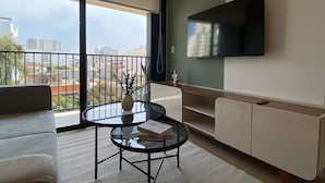 A modern living room with a glass coffee table, a sofa, a TV mounted on the wall, and a balcony with a view of buildings. - w Deluxe 2BR w Balcony View in Miraflores (Lima)