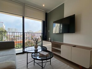 A modern living room with a glass coffee table, a sofa, a TV mounted on the wall, and a balcony with a view of buildings. - w Deluxe 2BR w Balcony View in Miraflores (Lima)