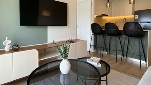 A modern living room with a flat-screen TV, a glass coffee table with a white vase, a sofa, and a kitchen area with bar stools. - w Deluxe 2BR w Balcony View in Miraflores (Lima)