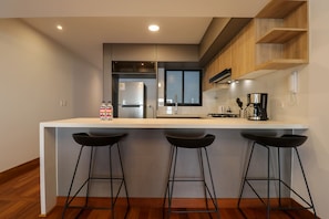 A modern kitchen with a white countertop, three black bar stools, a coffee machine, and wooden shelving. - w Homey 3BR With Balcony in Miraflores (Lima)