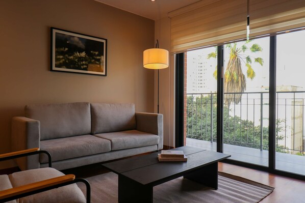 A modern living room with a sofa, a coffee table, a lamp, and a large window with a view of palm trees and buildings. - w Homey 3BR With Balcony in Miraflores (Lima)