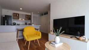 A modern living room with a yellow armchair, a round wooden table with books and a vase, a flat-screen TV, and a kitchen area with cabinets and a coffee machine. - w Luxury Cozy 2BR w Balcony in Miraflores (Lima)