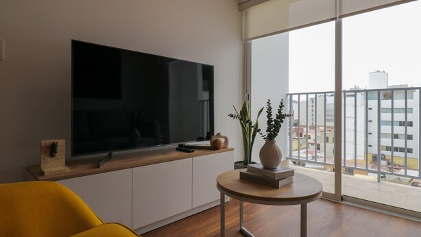 A modern living room with a flat-screen TV, a yellow armchair, a small round table with books and a vase, and a large window with a city view. - w Luxury Cozy 2BR w Balcony in Miraflores (Lima)