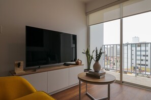 A modern living room with a flat-screen TV, a yellow armchair, a small round table with books and a vase, and a large window with a city view. - w Luxury Cozy 2BR w Balcony in Miraflores (Lima)