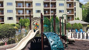 Un’area giochi con scivolo e strutture per arrampicarsi, circondata da una recinzione e con un edificio con diversi balconi sullo sfondo.