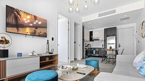 A modern living room with a TV, sofa, and ottomans, a dining area with a table and chairs, and a kitchen in the background. - NOK Luxury 1BR Apartment The Towers by Vistacana (Punta Cana)
