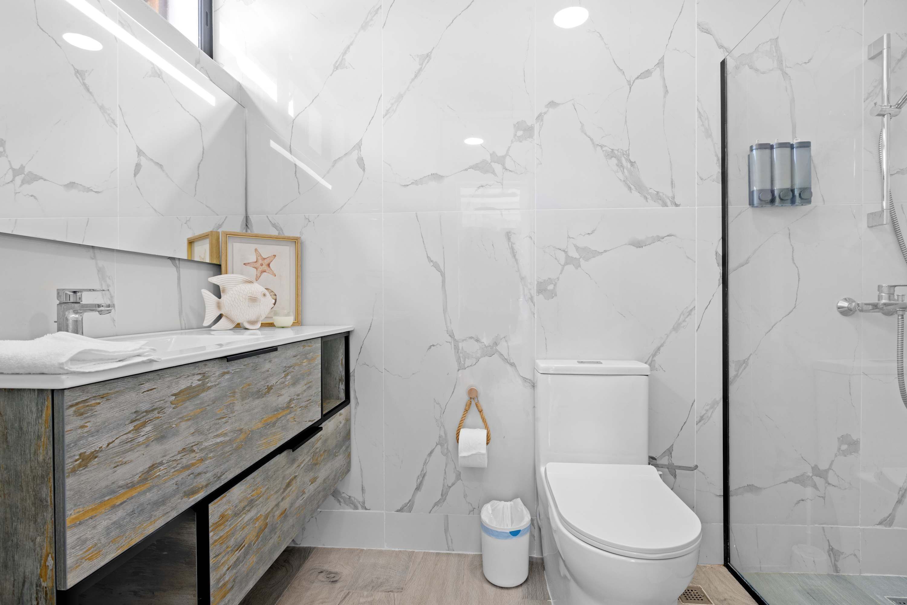 A bathroom with marble walls, a wooden vanity, a toilet, and a shower area.
