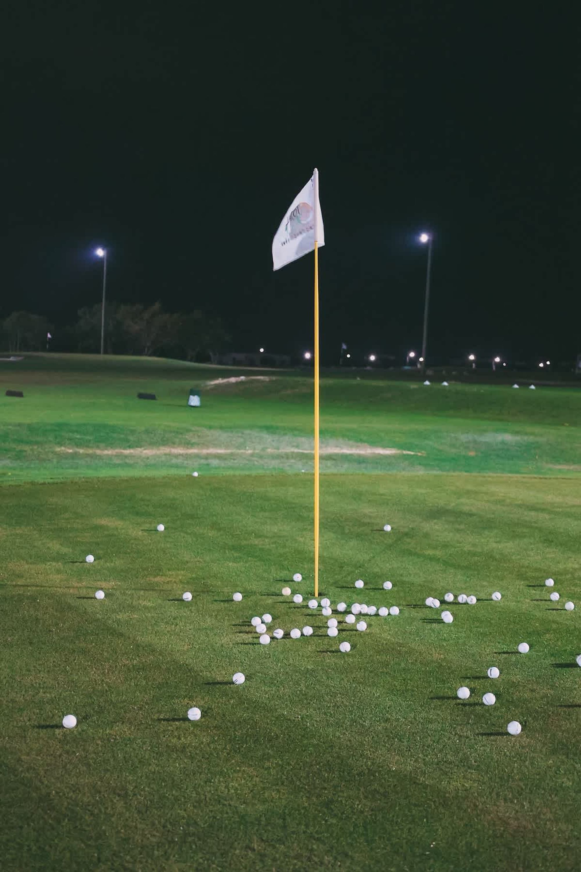 Ein Golfplatz bei Nacht mit einer Fahne auf dem Flaggenstock, beleuchtet durch Overhead-Leuchten, und verstreuten Golfbällen auf dem Rasen.