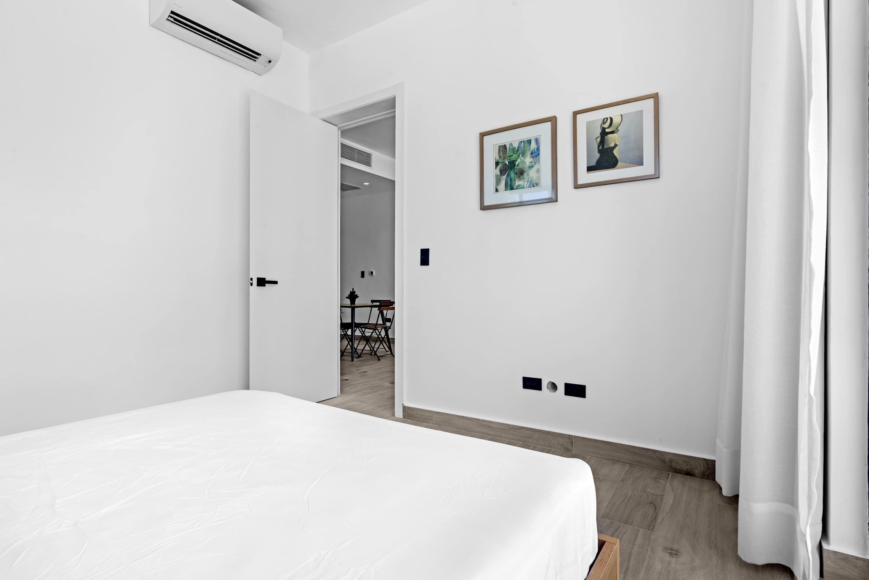 A modern hotel room with a bed, white walls, two framed artworks, and a door leading to a dining area.