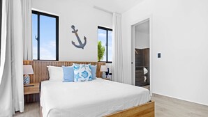 A bedroom with a bed, wooden headboard, white bedding, a nightstand with a lamp, a wall anchor decoration, and a view of the outside. - NOK Luxury 1BR Apartment The Towers by Vistacana (Punta Cana)