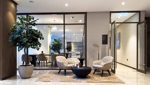 Eine moderne Lobby mit einer Sitzecke, einem großen Kübelbaum und einer Glaswand, die in einen Essbereich führt.