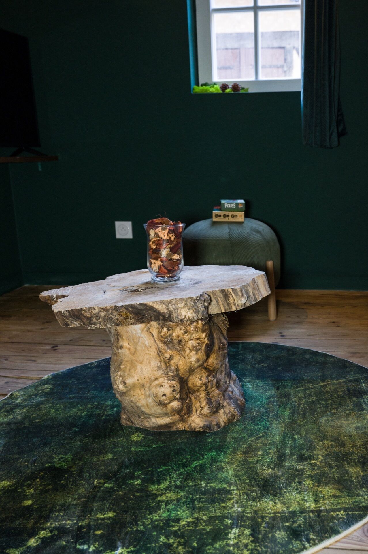 Een rustieke houten tafel met een glazen vaas op een groene vloerkleed, een raam met uitzicht, en een doosje met de tekst 'Pixies' op de achtergrond.
