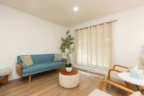 A living room with a blue sofa, a round coffee table, a small table with a tissue box, and a window with curtains. - Modern Renovated 4min Notredame 4bed 2bath 10peopl (South Bend)