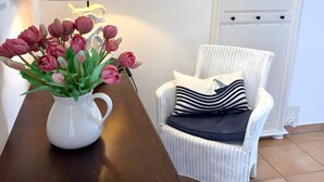 Ein weißer Korbstuhl mit gestreiftem Kissen, eine weiße Kanne mit rosa Tulpen und ein Holztisch.