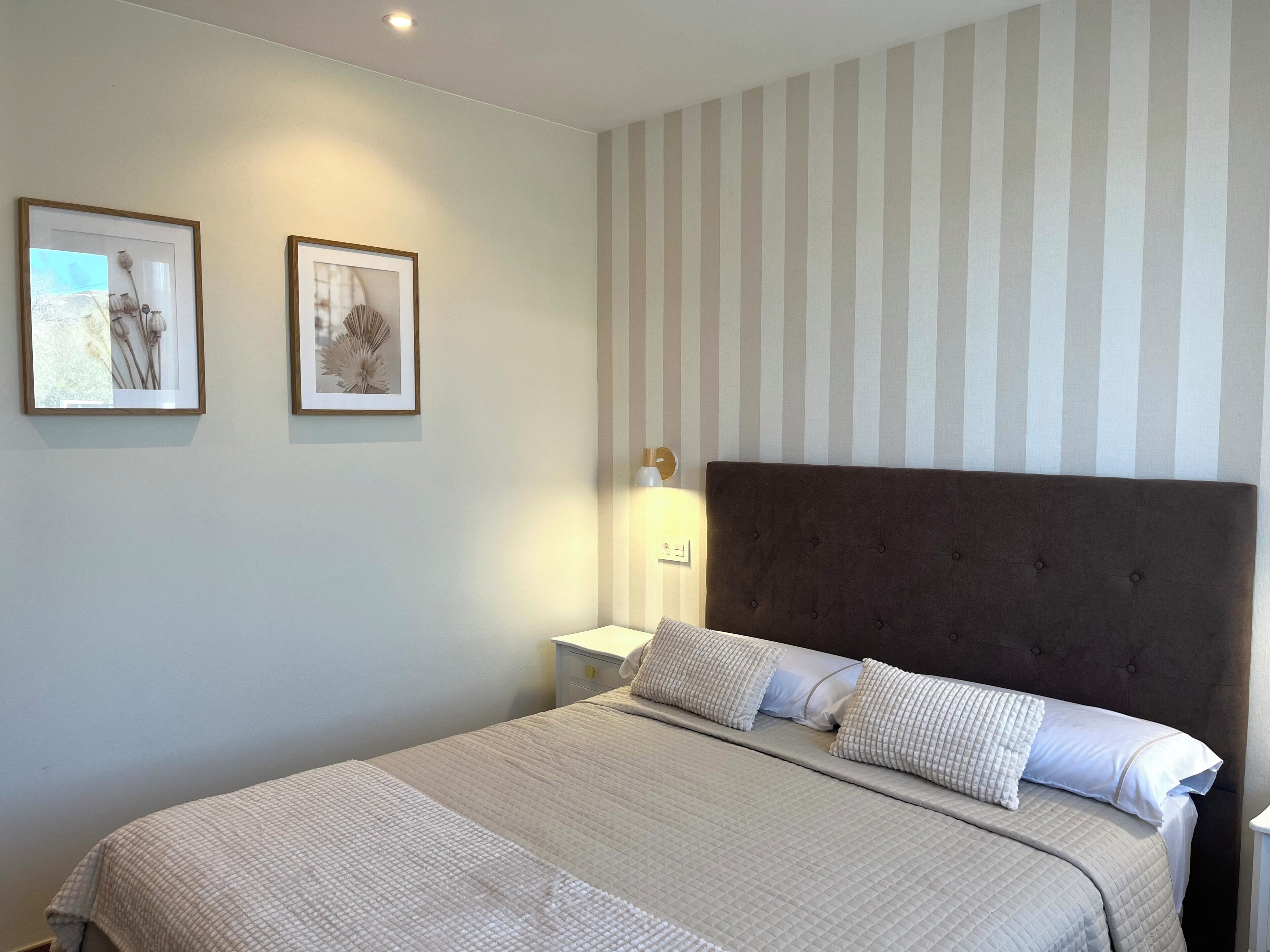 A bedroom with a bed, a nightstand, two framed pictures, a striped wall, and a wall-mounted lamp.