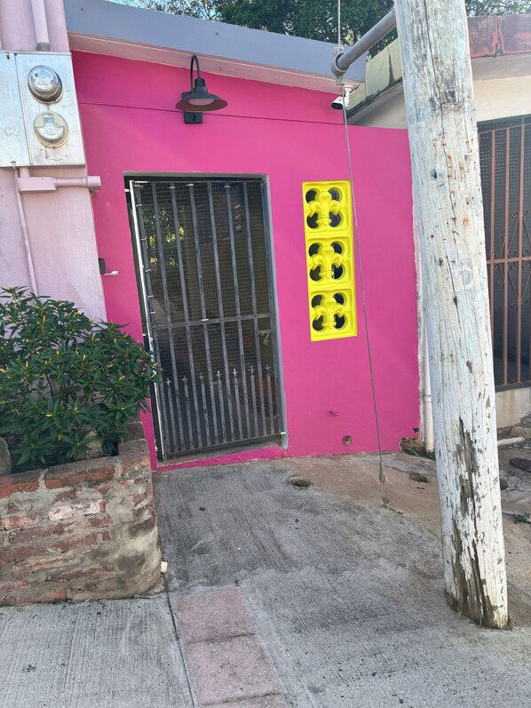 Un edificio rosa con una verja, un panel decorativo amarillo y una jardinera de ladrillo con follaje verde.