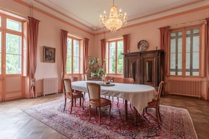 Een eetruimte met een ronde tafel, stoelen, een kroonluchter en grote ramen voorzien van gordijnen.
