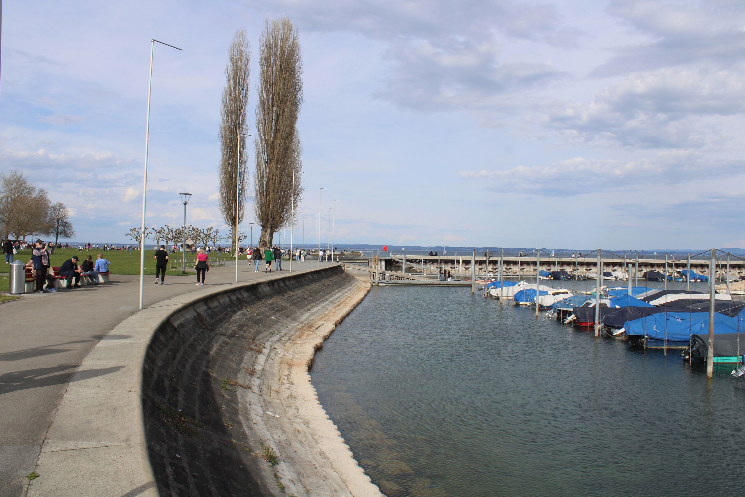 Eine Uferpromenade mit einer gewundenen Böschung, eine Reihe von Booten, die an der rechten Seite festgemacht sind, und Menschen, die den Weg entlanggehen.
