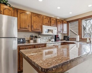 A kitchen with wooden cabinets, a stainless steel refrigerator, a microwave, and a granite countertop. - Farmstead Tee & Ski - Resort Core location, Fireplaces, Dog Friendly (Galena)