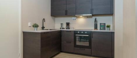 Cocina compacta con armarios oscuros, horno integrado y encimera con cafetera.