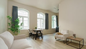 Un salón amplio con sofá, una mesa de comedor con sillas, una mesa de centro y ventanas grandes con cortinas.