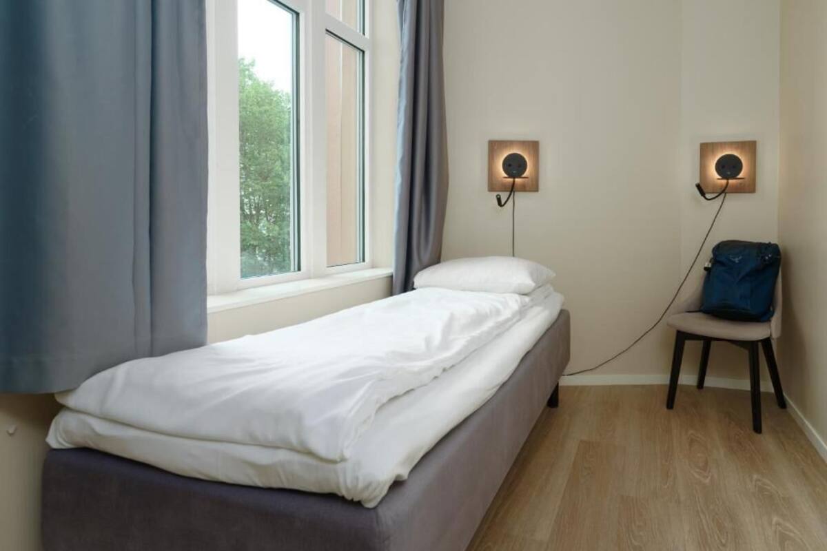 Ein Einzelbett mit weißer Bettwäsche, ein Sessel, eine Wandlampe und ein Fenster mit Vorhängen.