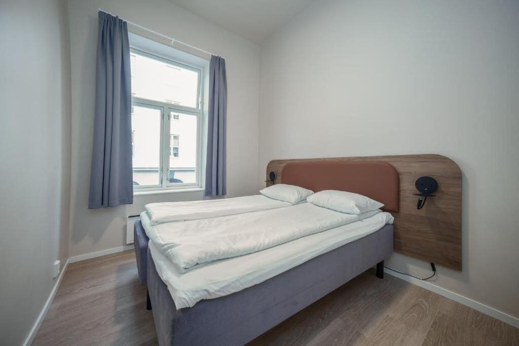 A single bed with a wooden headboard, a window with curtains, and a wall-mounted light fixture.